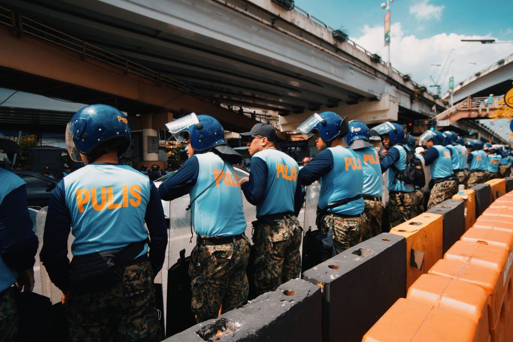 A line of police officers wearing helmets and blue vest labeled 'Pulis' during a daytime outdoor event under an overpass.