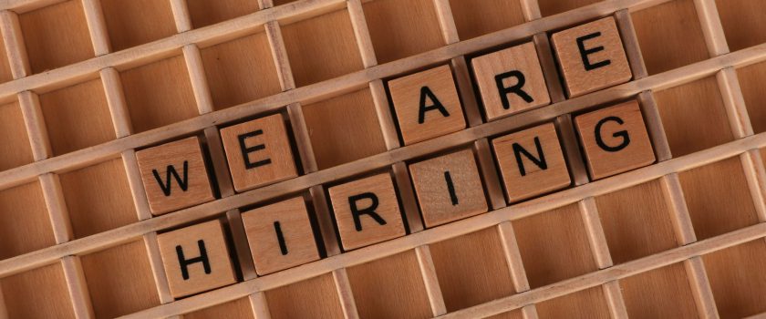 Wooden letter tiles arranged to spell 'We Are Hiring' on a wooden grid background.