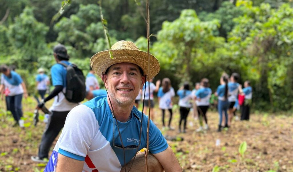 Andy Schachtel in a straw hat and Sourcefit shirt, smiling, with volunteers planting trees behind him