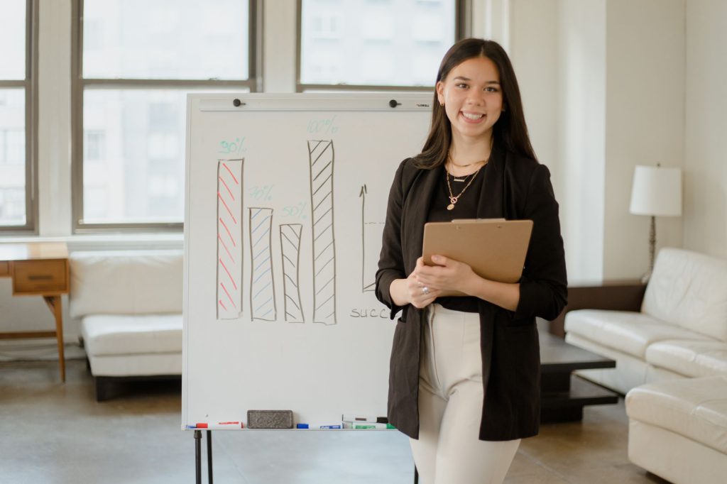 Confident businesswoman presenting charts and data analysis on a whiteboard in a modern office setting.
